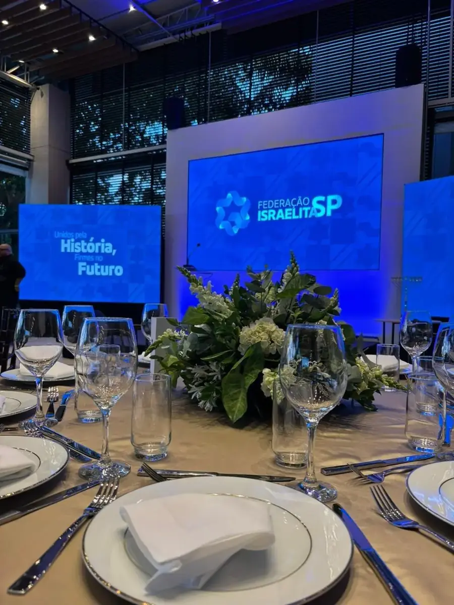 Mesa posta para evento corporativo institucional, com telões ao fundo