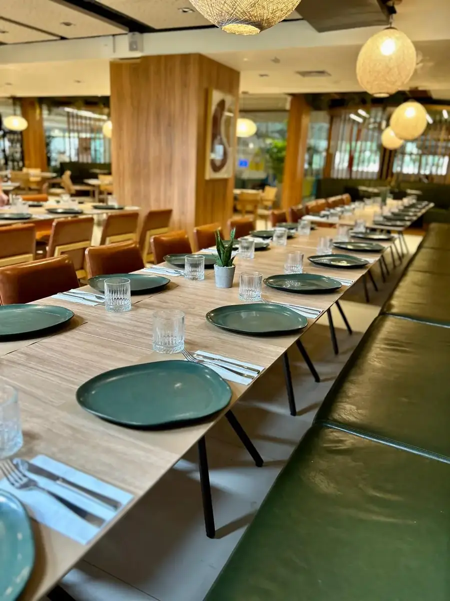 Mesa comprida posta no salão do restaurante, com pratos verdes e cadeiras de couro
