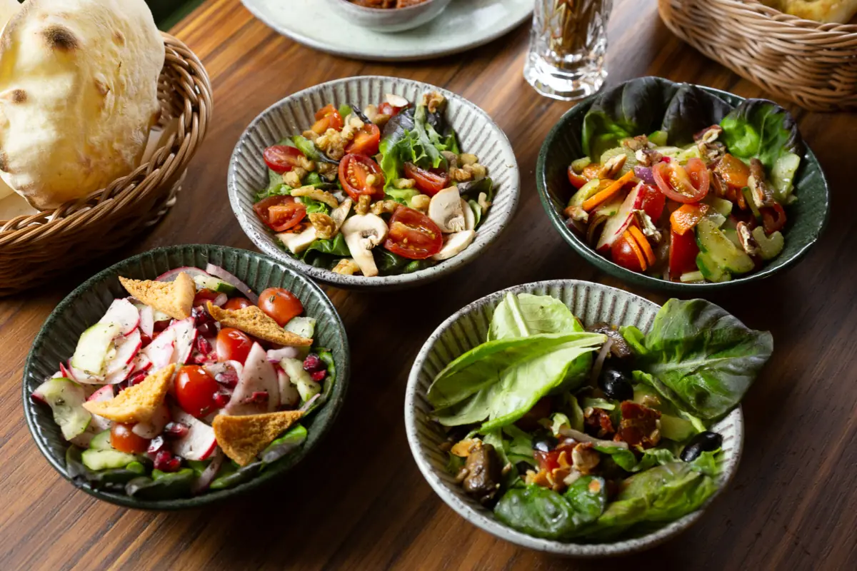Quatro saladas em tigelas de cerâmica — casa, fatuche, verde e arábias — com cesta de pitas